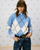 Model with her hands in her jeans pockets, standing against a wall with a small floral pattern. She wears a blue and cream argyle jumper with decorative stitching over a striped shirt. Her jeans feature Love Birds motifs and a black belt with a gold-tone buckle depicting the iconic PINKO birds. | Pinko