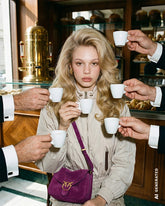 Blonde woman with wavy hair sitting in an Italian café, surrounded by hands offering her espresso cups, wearing a beige jacket and a purple suede crossbody bag with the Pinko logo. | Pinko