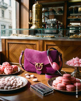 Fuchsia suede crossbody bag with the Pinko logo and pink headphones, placed on a table in an Italian café surrounded by macarons, meringues and pink pastries. | Pinko