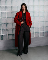 Model vor einer Glasbausteinwand, in einem langen roten Mantel über einem dunklen Anzug mit gegürtetem Blazer und weiter Hose. Es trägt schwarze spitze Schuhe und hält eine bordeauxrote Tasche unter dem Arm, verziert mit goldfarbenen Nieten und dem Love Birds Logo mit den ikonischen PINKO Vögeln. | Pinko