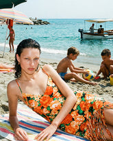 A woman lying on a striped beach towel wearing an orange floral mini dress with fringe detail, surrounded by a lively beach scene with children playing in the sand and the sea in the background. | Pinko