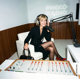 Blonde woman sitting in a radio studio with a "Pinko Radio" neon sign, wearing a black dress with a lace-up waist detail and red pumps, with headphones and a microphone, holding a black handbag. | Pinko
