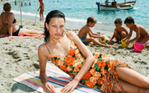 A woman lying on a striped beach towel wearing an orange floral mini dress with fringe detail, surrounded by a lively beach scene with children playing in the sand and the sea in the background. | Pinko