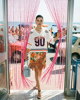 A woman wearing a white Pinko "Bold Soul 90" football t-shirt, an orange floral fringed mini skirt and black strappy heeled sandals, carrying a light blue Pinko bag, walking through a pink bead curtain with a retro beach scene in the background. | Pinko
