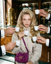 Blonde woman with wavy hair sitting in an Italian café, surrounded by hands offering her espresso cups, wearing a beige jacket and a purple suede crossbody bag with the Pinko logo. | Pinko