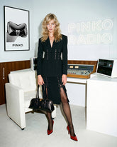AI-generated photoshoot featuring a model in a radio studio with a “PINKO RADIO” sign. She wears a black blazer and skirt with lace-up details, black tights and red pumps, and holds a glossy black handbag. | Pinko