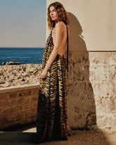 A woman wearing a backless tiger-print maxi dress poses against a sun-lit stone wall, with rocky coastline and blue sea in the background. | Pinko