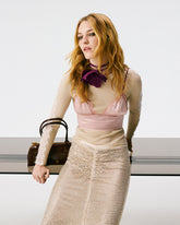 Eine Frau in einem cremefarbenen gerippten Langarmtop mit Spitzeneinsätzen, einem rosa Satin-Bralette, einem mit Kristallen verzierten Midi-Rock und einem lila Blumen-Choker, mit einer dunkelbraunen Pinko-Tasche im Bowling-Modell. | Pinko