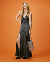 Model posing front-facing in a charcoal grey draped maxi dress with a deep V-neckline and front slit, paired with a fringed embellished handbag, set against a solid orange background. | Pinko