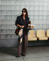 Model mit Sonnenbrille, dunklem Blazer und weiter Nadelstreifenhose vor einer Glasbausteinwand und gelben Sitzschalen. Es trägt eine bordeauxrote Umhängetasche mit Love Birds Logo und angeknotetem Tuch quer über dem Körper und hält ein Tablett mit To-go-Bechern und einer Papiertüte. | Pinko