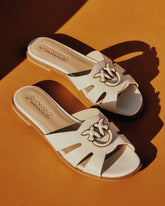 A pair of white leather Pinko flat slide sandals with cut-out strap detail and gold swallow logo hardware, on an orange surface. | Pinko