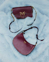 Zwei bordeauxrote Taschen mit goldfarbenen Nieten und dem Love Birds Logo mit den ikonischen PINKO Vögeln auf einer hellblauen Oberfläche in Felloptik. Oben eine rechteckige Tasche mit Überschlag und einem Riemen mit Kettenabschnitten, darunter eine halbmondförmige Tasche mit schmalem Riemen. | Pinko