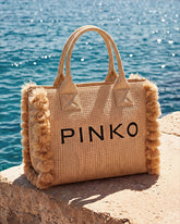 A large natural woven raffia Pinko beach tote bag with fluffy pom-pom trim on the sides, placed on a stone surface by the sea. | Pinko