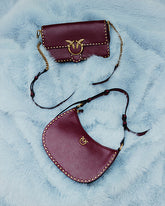 Zwei bordeauxrote Taschen mit goldfarbenen Nieten und dem Love Birds Logo mit den ikonischen PINKO Vögeln auf einer hellblauen Oberfläche in Felloptik. Oben eine rechteckige Tasche mit Überschlag und einem Riemen mit Kettenabschnitten, darunter eine halbmondförmige Tasche mit schmalem Riemen. | Pinko