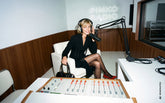 Blonde woman sitting in a radio studio with a "Pinko Radio" neon sign, wearing a black dress with a lace-up waist detail and red pumps, with headphones and a microphone, holding a black handbag. | Pinko